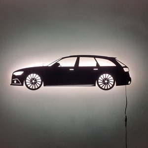 May include: A black silhouette of a station wagon car with a light shining through it. The car is on a white wall.
