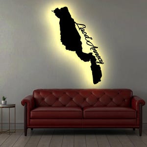 May include: A black silhouette of a lake with the text "Lac d'Annecy" is mounted on a wall with a warm yellow glow. The wall art is above a red leather sofa. A chair, side table, and potted plant complete the room.