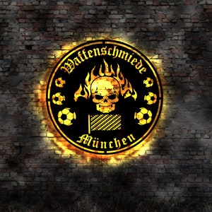 Puede incluir: Un logotipo circular negro y dorado con el texto "Waffenschmiede München" y una calavera con llamas detrás. El logotipo está sobre un fondo de pared de ladrillo.