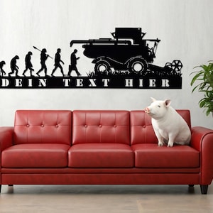 May include: Black metal wall art showing the evolution of man to a combine harvester, with the text "DEIN TEXT HIER". A red leather sofa and a white pig are in the foreground. A potted plant is on the right.