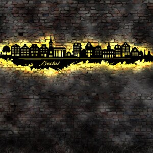 May include: A black metal silhouette of the city skyline of Liestal, Switzerland, with the city name written below. The silhouette is lit from behind with a warm yellow light, creating a glow effect. The silhouette is mounted on a brick wall.