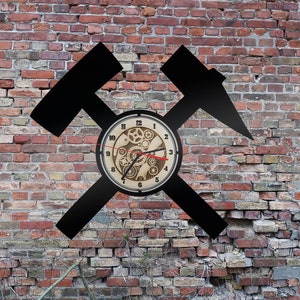 May include: A black metal clock with a hammer and pickaxe design. The clock face is made of wood and features a gear design. The clock is mounted on a brick wall.