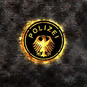 Op de afbeelding: Een cirkelvormig embleem met het woord "POLIZEI" in witte letters, met een gouden adelaar, tegen een zwarte achtergrond. Het embleem wordt verlicht door een vurige gloed, gemonteerd op een verweerde bakstenen muur.