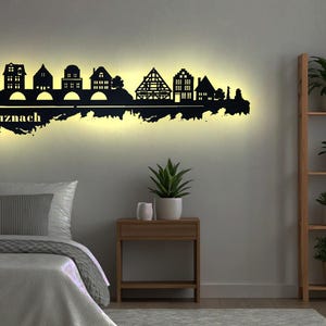 May include: A black metal wall art silhouette of the Bad Kreuznach skyline, illuminated with a warm glow. The artwork features detailed building outlines and the city name. The piece is mounted above a bed in a bedroom setting.