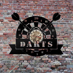 May include: A black metal wall clock shaped like a dartboard with a clock face in the center. The clock face has a gear design and the words "DARTS" are printed below the clock face.