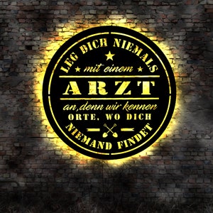 Könnte beinhalten: Ein schwarzes und gelbes rundes Schild mit dem Text "LEG DICH NIEMALS mit einem ARZT an, denn wir kennen ORTE, WO DICH NIEMAND FINDET" auf einem Ziegelmauer Hintergrund.