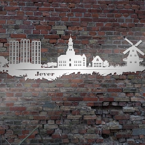 May include: White metal silhouette of the Jever skyline, featuring buildings, a windmill, and the word "Jever" on a brick wall background.