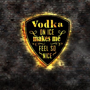 May include: A black shield-shaped sign with the words "Vodka ON ICE makes me FEEL SO NICE" in yellow lettering. The sign has a glowing orange border and is mounted on a brick wall.
