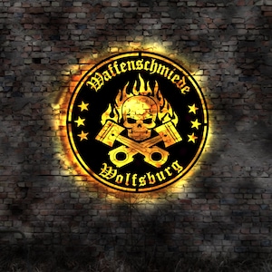 Può includere: Un cartello circolare nero e dorato con il testo "Waffenschmiede Wolfsburg" e un teschio con fiamme dietro.