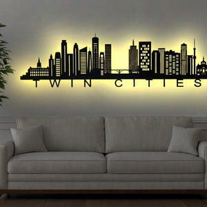 以下が含まれることがあります： 背面から照らされた「TWIN CITIES」の文字が入った、都市のスカイラインを描いた黒い金属製のウォールアート。アートは灰色のソファの上に設置され、フロアランプと鉢植えの木が両側にあります。