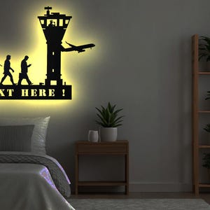 Op de afbeelding: Zwarte metalen wandkunst die de evolutie van de mensheid afbeeldt met een toren en een vliegtuig. De kunst is verlicht met gele lampen en heeft de tekst "YOUR TEXT HERE!" eronder.