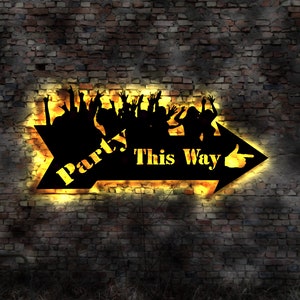 Könnte beinhalten: Ein schwarzes Pfeilschild mit den Worten "Party This Way" und Silhouetten von tanzenden Menschen. Das Schild ist mit einem warmen gelben Schein beleuchtet und an einer Backsteinwand befestigt.