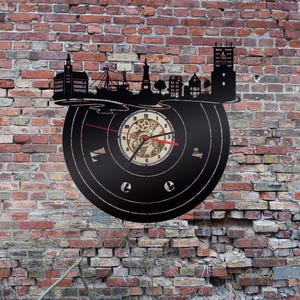 May include: A black vinyl record clock featuring a cityscape silhouette and a clock face with gears. The clock is mounted on a brick wall.