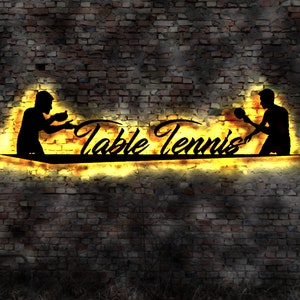 Puede incluir: Un letrero de neón con las palabras "Table Tennis" en una fuente estilizada. Dos siluetas de personas jugando al tenis de mesa están a cada lado del texto. El letrero está iluminado con un cálido resplandor amarillo.