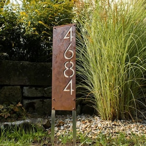 May include: A rusty metal house number sign with the numbers 4684 cut out. The sign is mounted on two metal stakes and is displayed in a garden setting.