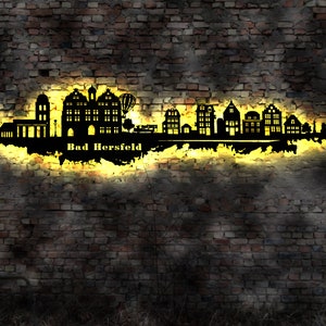 May include: A black silhouette of a cityscape with the text "Bad Hersfeld" below it. The silhouette is illuminated with a yellow glow against a brick wall background.