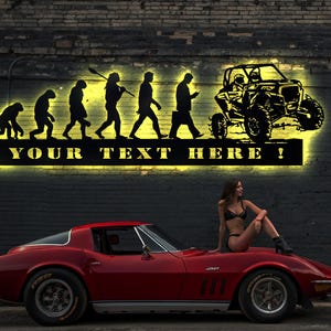 May include: A red sports car with a woman sitting on the hood. The car is parked in front of a brick wall with a metal sign that says "YOUR TEXT HERE!" The sign features a silhouette of the evolution of man, ending with a person driving a side-by-side off-road vehicle.