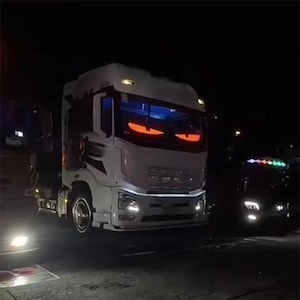 Puede incluir: Un camión blanco con ojos rojos en el parabrisas delantero. El camión está aparcado de noche y las luces delanteras están encendidas.