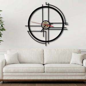 May include: A black metal wall clock with a geometric design. The clock has a round face with a smaller circle in the centre. The clock is hanging on a white wall.