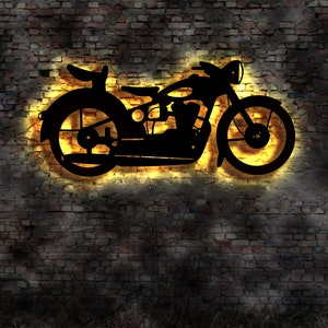 May include: A black metal silhouette of a vintage motorcycle with a yellow glow behind it, mounted on a brick wall.