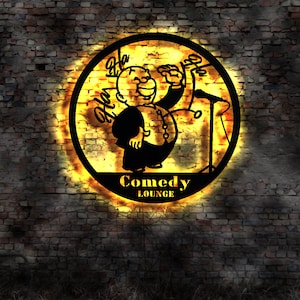 Puede incluir: Un letrero de metal negro y amarillo con una imagen de dibujos animados de un hombre riendo y el texto "Comedy Lounge".
