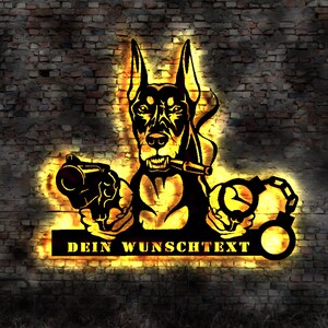 Könnte beinhalten: Schwarze Metall-Wandkunst mit einem Dobermann mit Zigarre, der eine Pistole und Handschellen hält. Der Text "DEIN WUNSCHTEXT" befindet sich unter dem Hund. Die Kunst wird von einem warmen, gelben Schein vor einer Backsteinmauer beleuchtet.