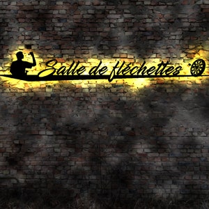 Puede incluir: Un letrero de metal negro con las palabras "Salle de fléchettes" en una fuente cursiva, iluminado por un resplandor amarillo. El letrero presenta una silueta de una persona lanzando un dardo y una diana. El letrero está montado en una pared de ladrillo.
