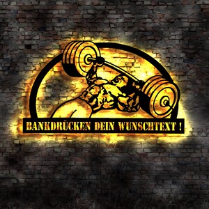 Może przedstawiać: Czarny i złoty metalowy znak przedstawiający sztangistę podnoszącego sztangę. Znak jest umieszczony na tle ceglanej ściany z napisem "BANKDRÜCKEN DEIN WUNSCHTEXT!" w prostokątnym polu.