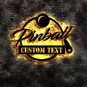 May include: A vintage-style neon sign with a glowing yellow circle and the word "Pinball" in black script. The sign also has a black and white image of a pinball and the text "CUSTOM TEXT" in a black rectangle.