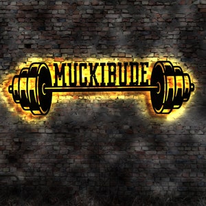 Peut inclure: Une barre d'haltères noire avec le mot "MUCKIRIDE" en lettres blanches, sur un mur de briques avec une lueur jaune autour de la barre.