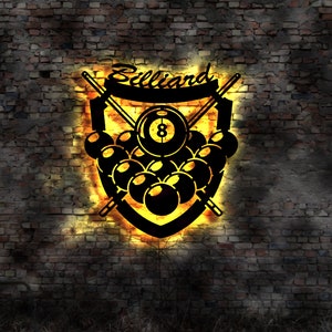 Könnte beinhalten: Ein schwarzes Billard-Metallschild mit einem Schilddesign, das gekreuzte Queues, Billardkugeln und die Zahl 8 zeigt. Das Wort "Billiard" ist oben gewölbt. Das Schild wird von einem warmen, gelben Schein vor einer Backsteinmauer beleuchtet.