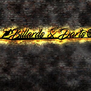 Könnte beinhalten: Ein schwarzes Metallschild mit den Worten "Billiards & Darts" in einer stilisierten Schrift. Das Schild ist mit warmen gelben Lichtern beleuchtet und an einer Backsteinwand montiert.