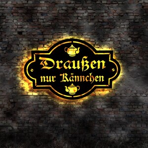 Puede incluir: Un letrero de metal negro con un contorno dorado y un brillo amarillo. El letrero dice "Draußen nur Kännchen", que se traduce como "Solo teteras afuera".