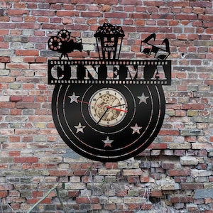 Könnte beinhalten: Schwarze Wanduhr zum Thema Kino. Die Uhr zeigt ein Design mit Filmrolle, Popcorn und 3D-Brille. Das Wort "CINEMA" wird über dem Zifferblatt angezeigt. Die Zeiger der Uhr sind rot, und die Uhr befindet sich vor einer Backsteinmauer.