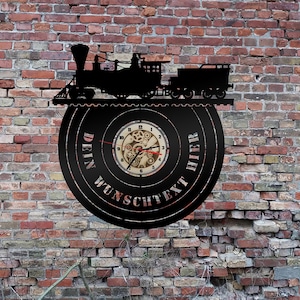 Puede incluir: Un reloj de metal negro con una silueta de una locomotora y una esfera de reloj con engranajes. La esfera del reloj tiene el texto "Dein Wunschtext Hier" alrededor de la esfera.