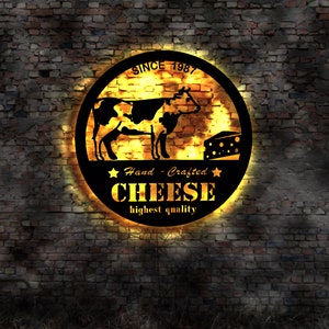 Könnte beinhalten: Ein schwarzes und gelbes rundes Schild mit dem Text "SINCE 1987" und "Hand-Crafted CHEESE highest quality" auf einem Hintergrund aus Backsteinmauer. Auf dem Schild sind auch eine schwarze Silhouette einer Kuh und ein Stück Käse abgebildet.