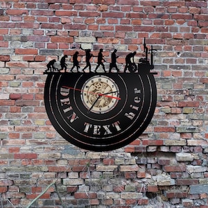 May include: A black vinyl record clock with a clock face featuring a gear design. The clock is mounted on a brick wall. The clock has the text "DEIN TEXT HIER" around the clock face.