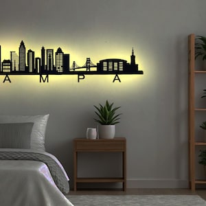 May include: A black metal wall art silhouette of the Tampa skyline, illuminated with warm lighting. The word "TAMPA" is spelled out below the skyline. The art is mounted above a bed in a bedroom.