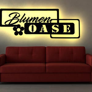 Könnte beinhalten: Ein schwarzes Wandzeichen aus Metall mit den Worten "Blumen Oase" und einem stilisierten Blumendesign. Das Schild ist mit warmweißen Lichtern beleuchtet.