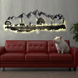 May include: A black metal wall art piece showing a mountain range and a rural scene with animals and people, lit by a warm light. A red leather sofa and a pig are in the foreground.
