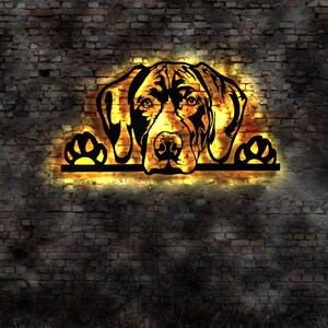 May include: A black metal silhouette of a dog's head with paws on either side, illuminated with a warm yellow glow against a brick wall background.