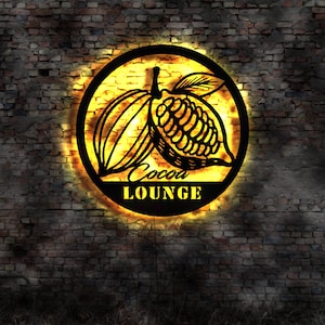 Könnte beinhalten: Ein schwarzes Metallschild mit einem gelben Schein dahinter. Das Schild zeigt die Silhouette von zwei Kakaobohnen und den Text "Cocoa Lounge".