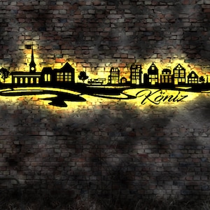 May include: A black metal silhouette of a town skyline with the word "Köniz" in a cursive font. The silhouette is illuminated with a yellow glow, mounted on a brick wall. The design features buildings, trees, and a church.