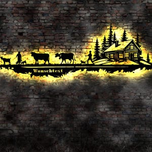 May include: Black metal silhouette of a cabin in the woods with a person walking a dog, two cows, and a goat. The silhouette is illuminated with a yellow glow and the text "Wunschtext" is below the silhouette.