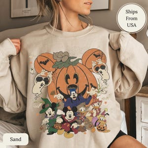 May include: A sand-colored crewneck sweatshirt featuring a large graphic print of Mickey Mouse and friends in Halloween costumes. The design includes a central orange pumpkin, ghosts, bats, and characters dressed as witches and vampires in a vintage style.