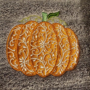 Puede incluir: Una calabaza naranja decorativa con un diseño floral blanco intrincado. La calabaza está bordada en una tela gris y peluda.
