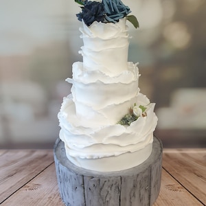 May include: A four-tier white wedding cake with ruffled frosting and blue and white flowers on top. The cake is on a grey wooden cake stand.