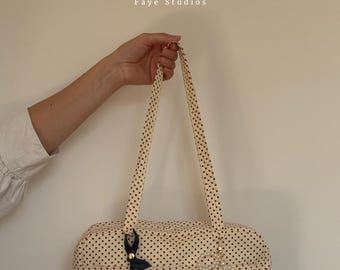 May include: A cream-colored mini bowler bag with a black polka dot pattern, featuring two long shoulder straps. The bag is adorned with a dark cherry charm and a white pearl-beaded bow charm. Text reads: The Mini Bowler Bag Pattern Faye Studios.