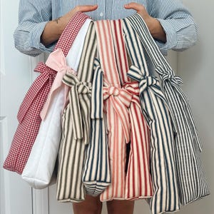 Op de afbeelding: Een verzameling stoffen draagtassen in verschillende patronen en kleuren. De tassen bevatten rood geruite, witte, roze en gestreepte ontwerpen in rood, marineblauw en zwart. Elke tas is vastgebonden met een bijpassende strik.