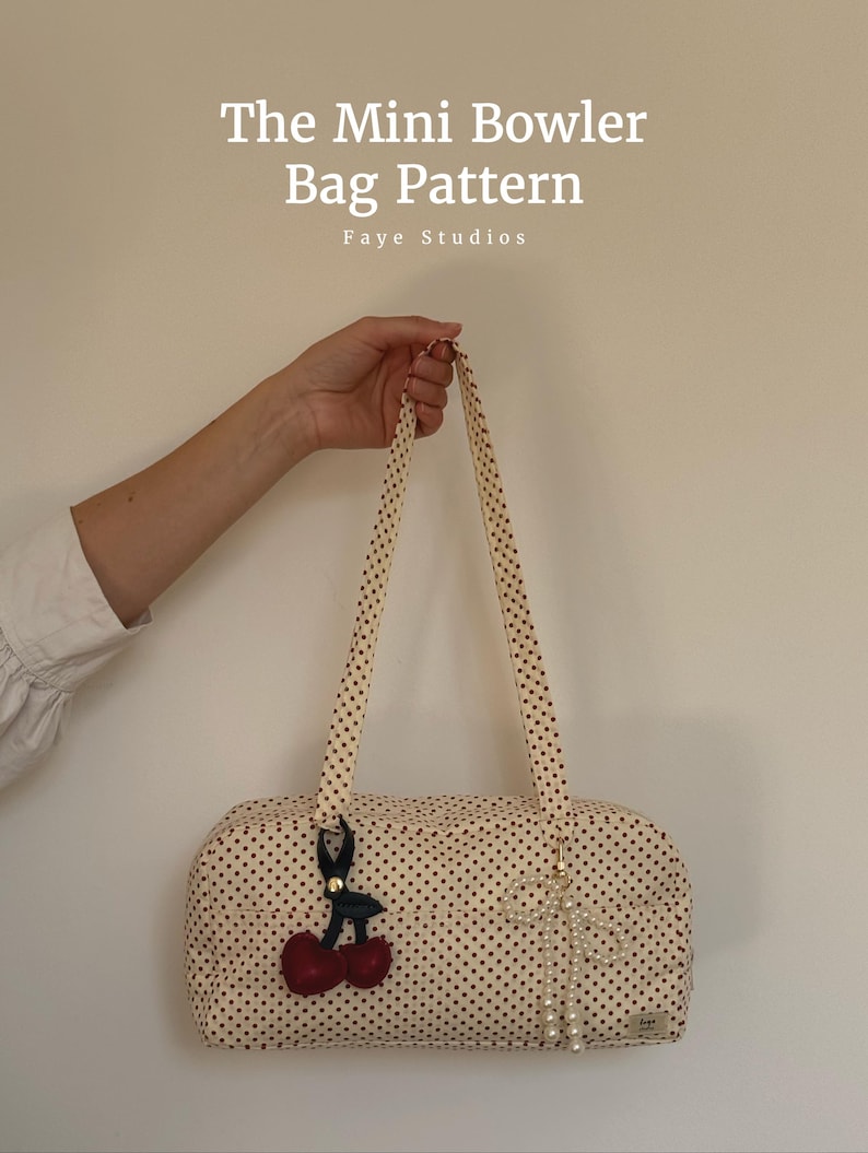 May include: A cream-coloured mini bowler bag with a black polka dot pattern, featuring two long shoulder straps. The bag is adorned with a dark cherry charm and a white pearl-beaded bow charm. Text reads: The Mini Bowler Bag Pattern Faye Studios.